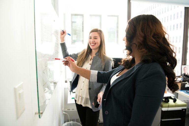 Two smiling, professional women collaborate at a whiteboard