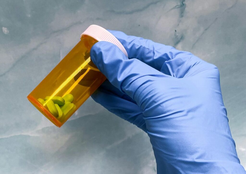 A hand wearing a blue latex glove holds a prescription bottle with a dozen or so large pills